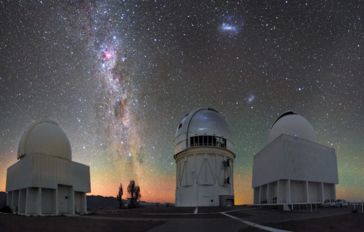 The stars and dust lanes of the Milky Way hang above telescope domes in this richly hued image of Cerro Tololo Inter-American Observatory (CTIO), a program of NSF’s NOIRLab. The two bright, wispy ovals to the right are the Large and Small Magellanic Clouds, dwarf galaxies visible only in the southern hemisphere. The band of color along the horizon is a phenomenon known as airglow, a visible reaction between molecules in Earth’s atmosphere and solar radiation. In this picture are the Curtis Schmidt Telescope (left), the Víctor M. Blanco 4-meter Telescope (middle), and the SMARTS 1.5-meter Telescope (right). These telescopes, alongside 12 others, comprise CTIO, the principal ground-based optical platform for US astronomical investigation of the southern skies.