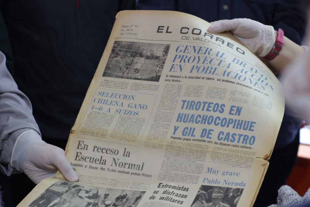Un periódico antiguo tomado por dos manos con guantes. El periódico se llama El Correo de Valdivia.